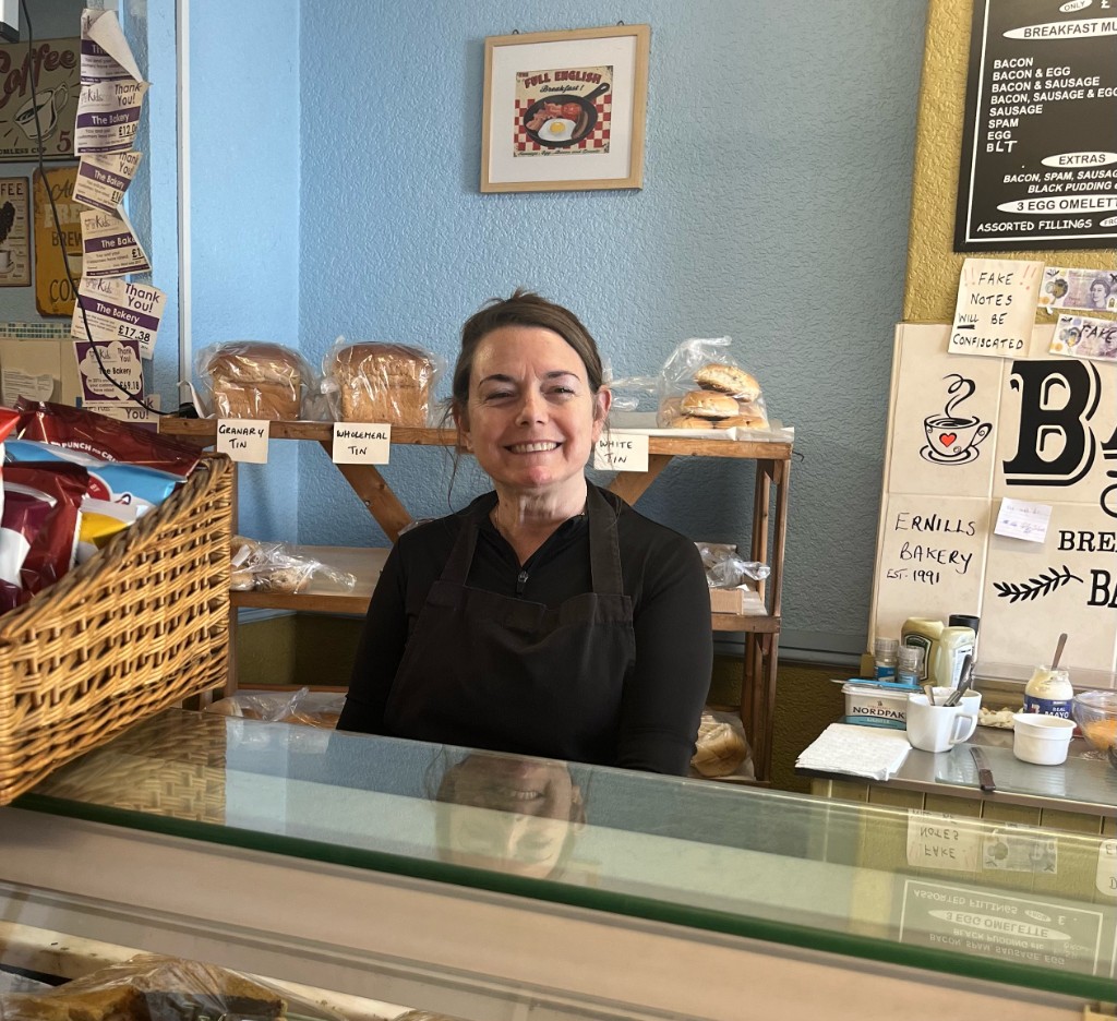 Stella, the owner of The Bakery Cafe Heywood, smiling behind the counter with fresh loaves of Granary and Wholemeal bread on the shelves behind her.